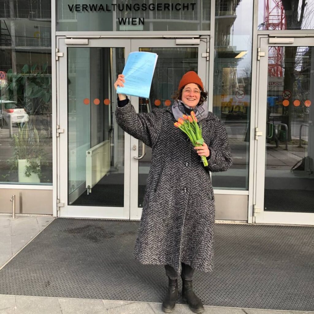LINKS Sprecherin Angelika Adensamer vor dem Gerichtsgebäude mit einem Blumenstrauß in der Hand. Sie lächelt in die Kamera und hält in der anderen Hand freudig eine Mappe in die Luft.